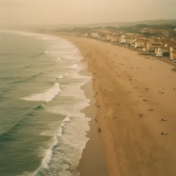 An aerial view of a long sandy beach with waves, people, and a coastal town.