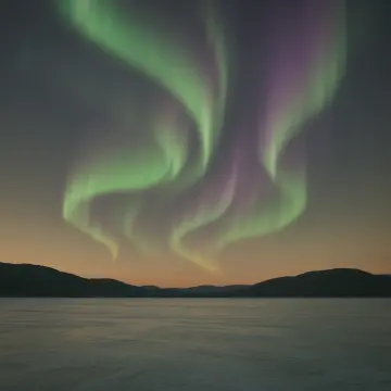 Green and purple aurora borealis illuminates the night sky above a frozen lake and distant mountains.