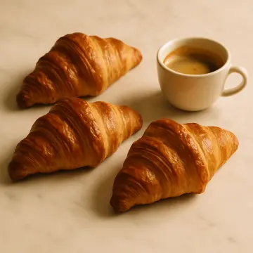 Three golden-brown croissants are arranged around a white cup of espresso on a light surface.