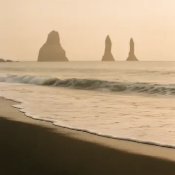 A misty morning at a dark sand beach with waves and distant sea stacks.