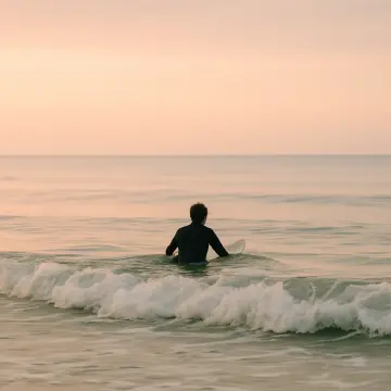 A surfer in a wetsuit sits on a surfboard in calm ocean water at dawn.